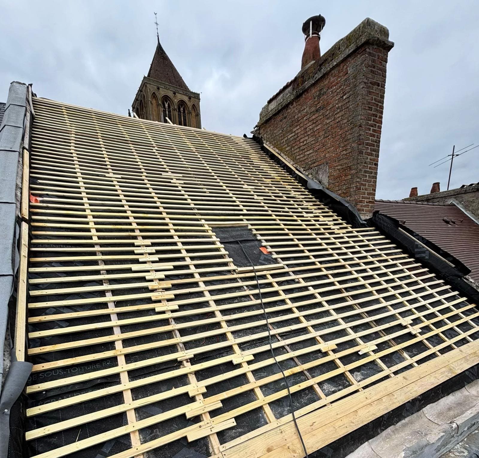 Liteaux en bois installés sur une toiture en rénovation avec un clocher en arrière-plan.