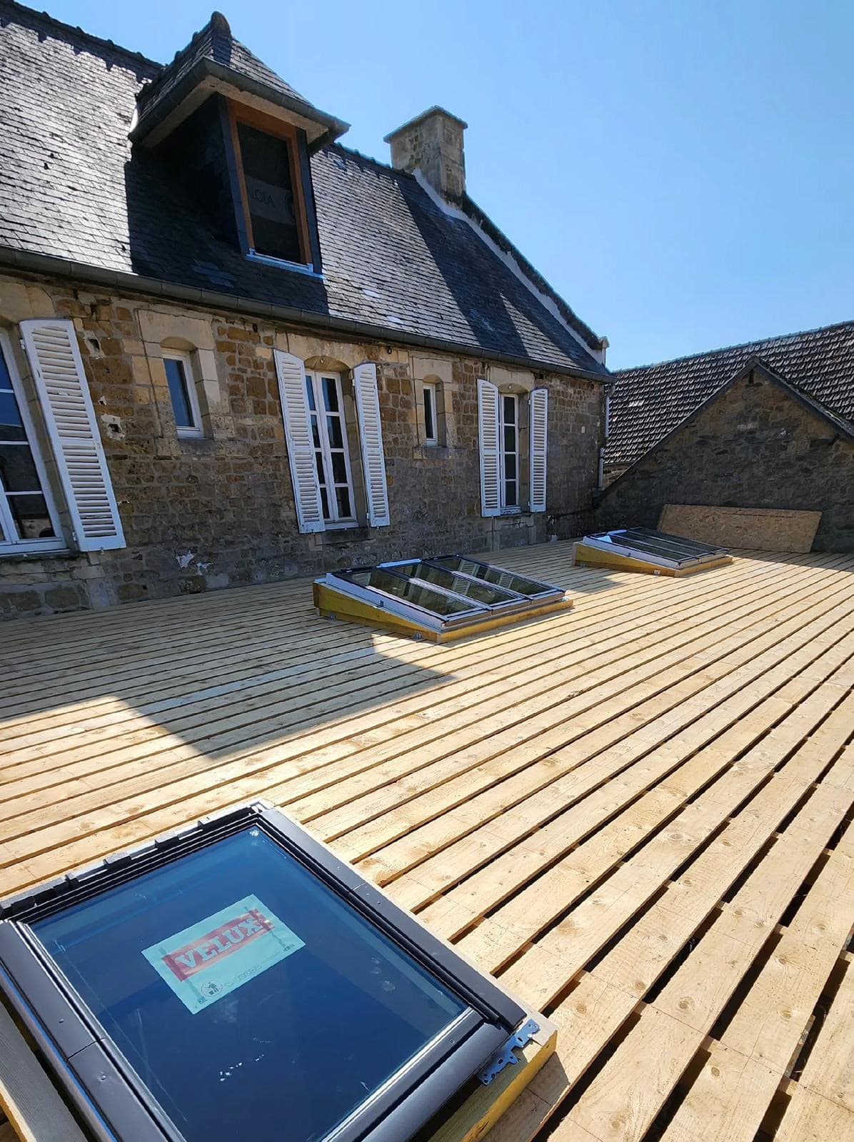 Fenêtres de toit Velux intégrées dans une terrasse en bois clair devant une maison en pierre.
